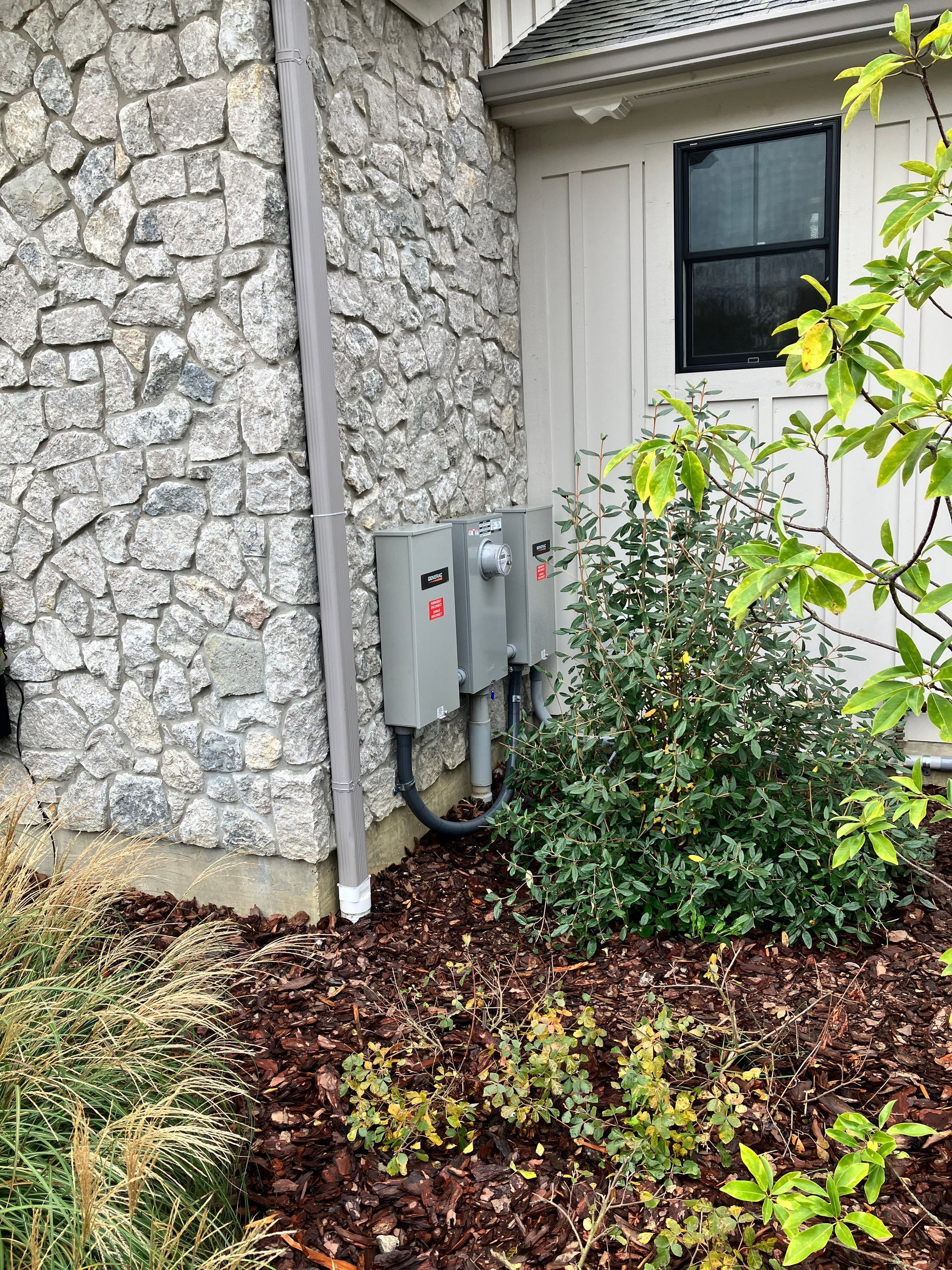 Electrical boxes on stone wall next to a bush and downspout. Landscaped yard with brown mulch.