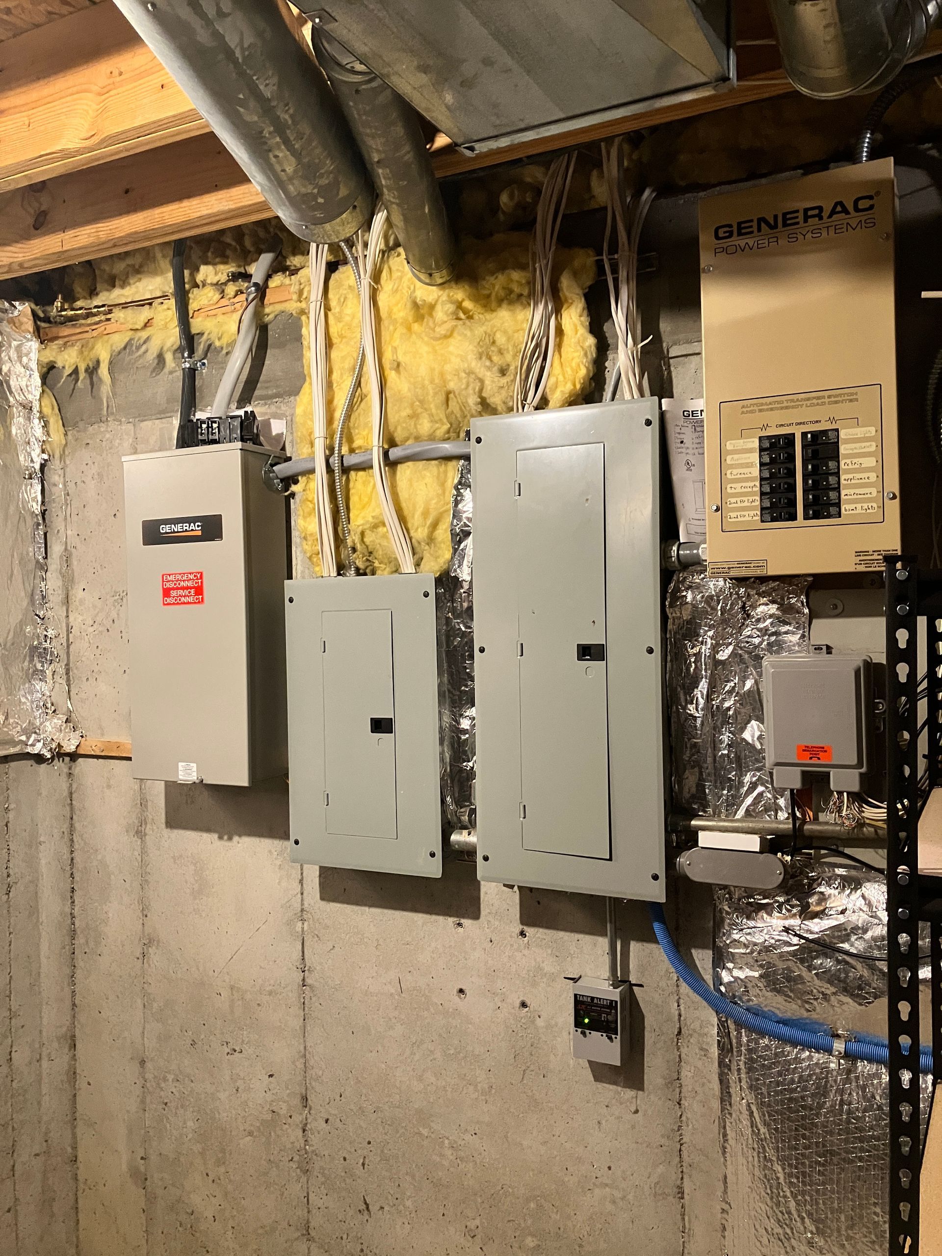 Electrical panels mounted on a concrete basement wall, with wiring and insulation visible.