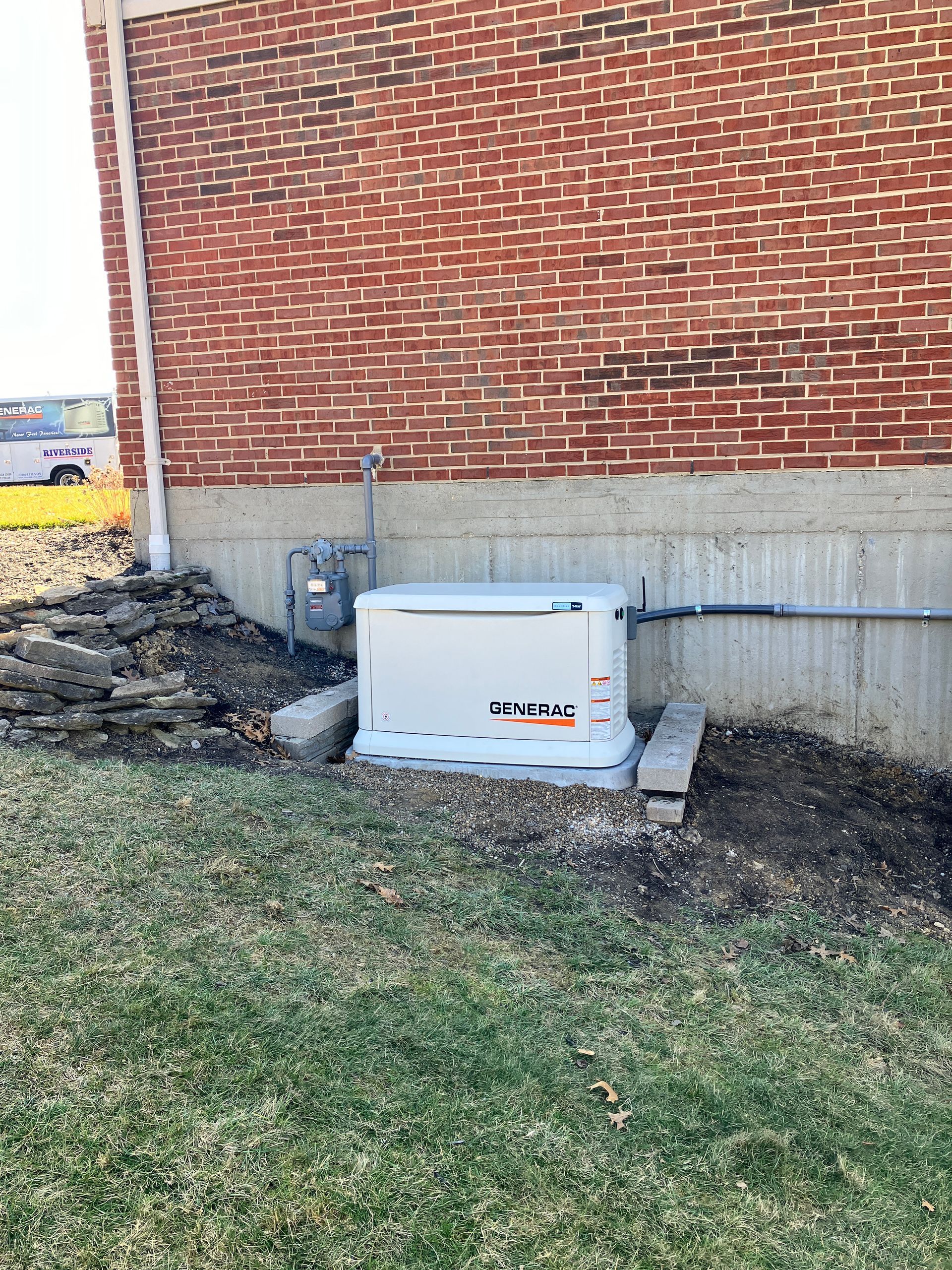 Generator unit next to a brick building. Ground is grass, with a pile of rocks on the left.