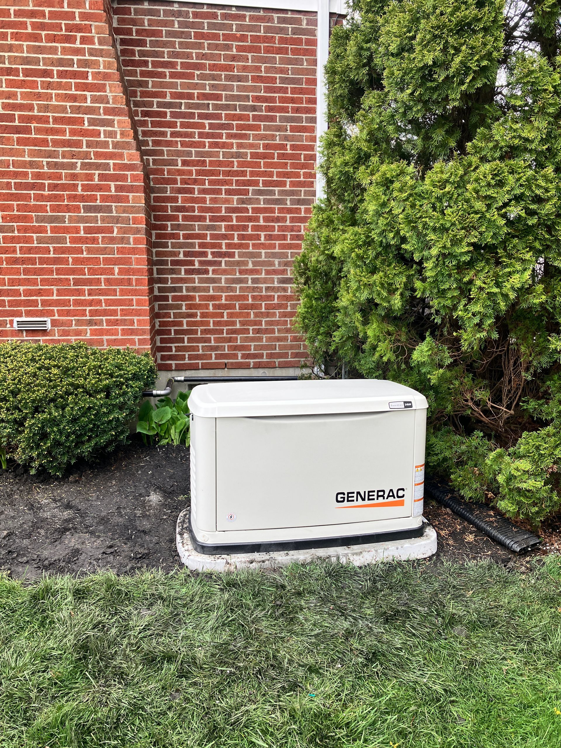 White generator on concrete pad next to a brick wall and bushes in a yard.