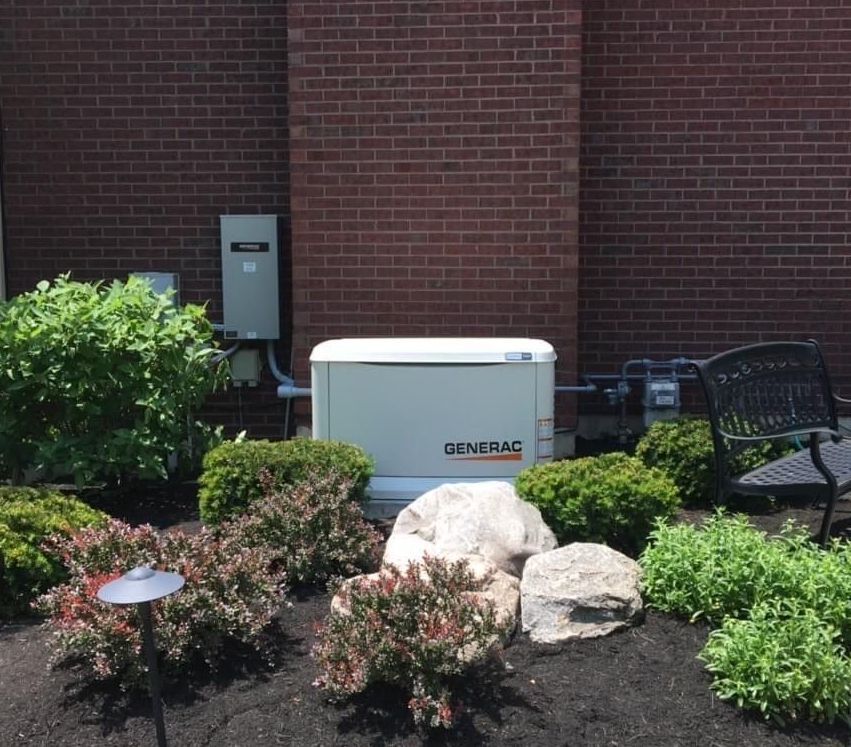 A Generac generator in a landscaped bed against a brick wall.
