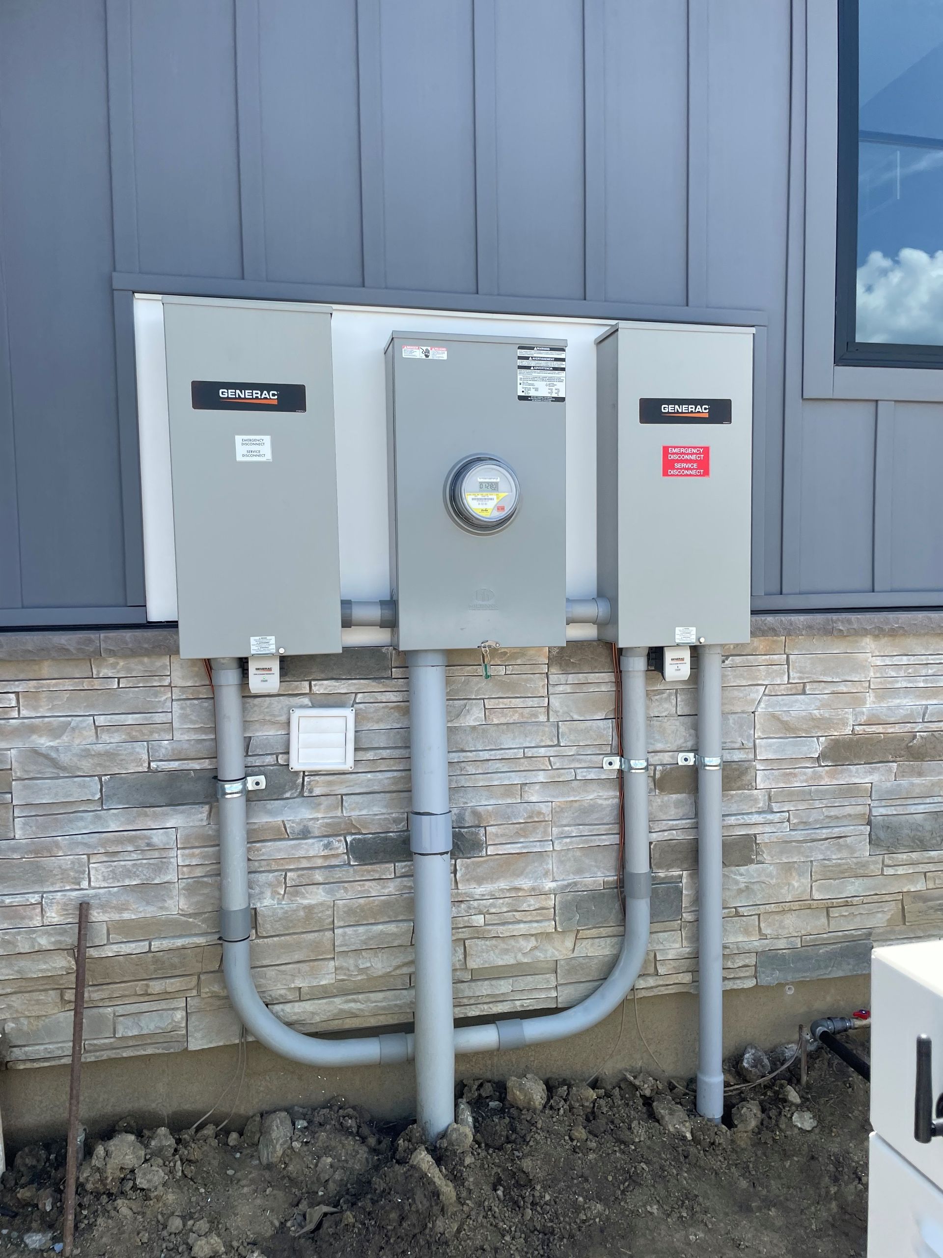 Exterior electrical panel with conduit against a gray siding wall and stone base.