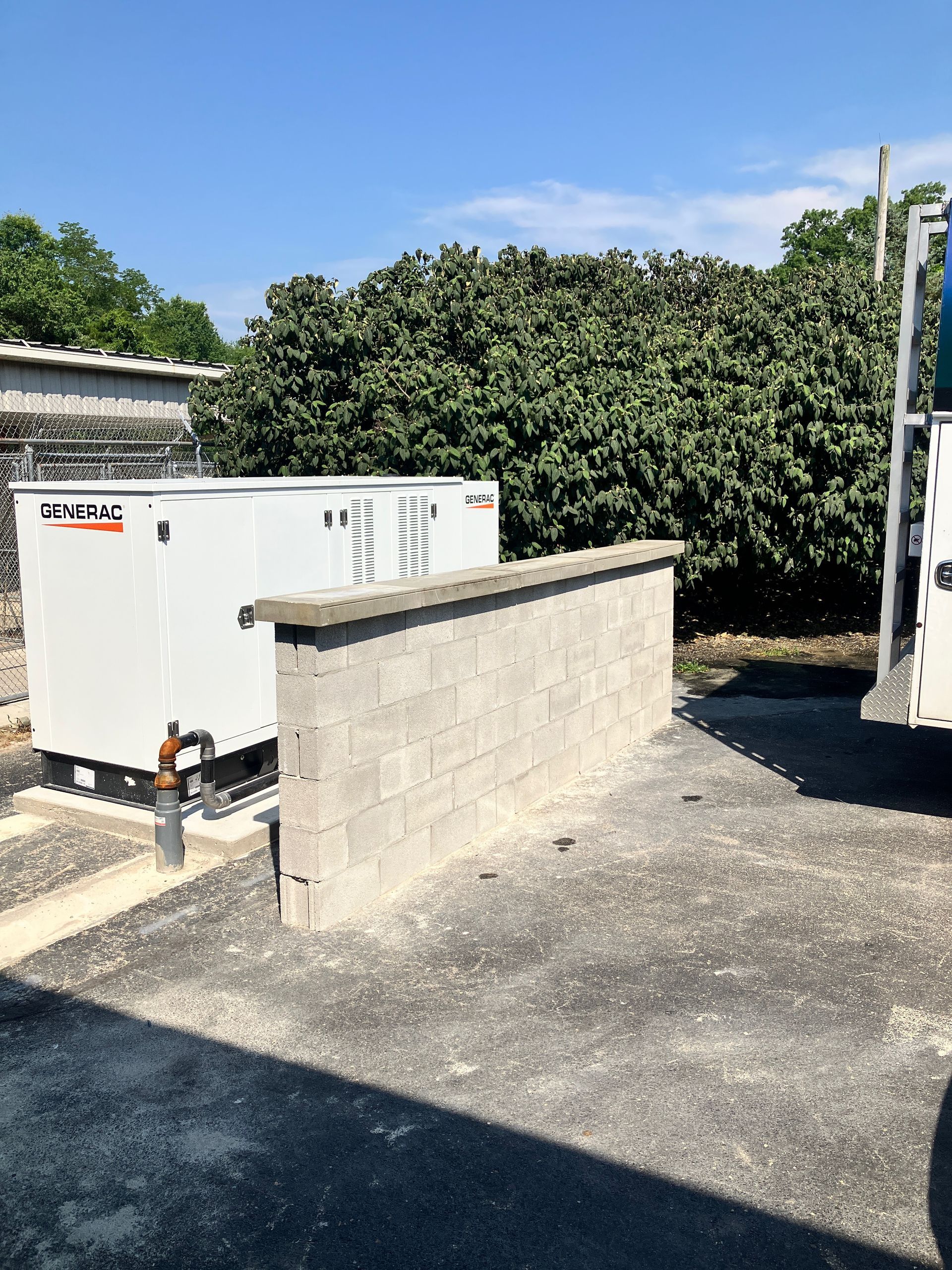 Generator behind a cinder block wall; surrounded by asphalt and trees under a blue sky.