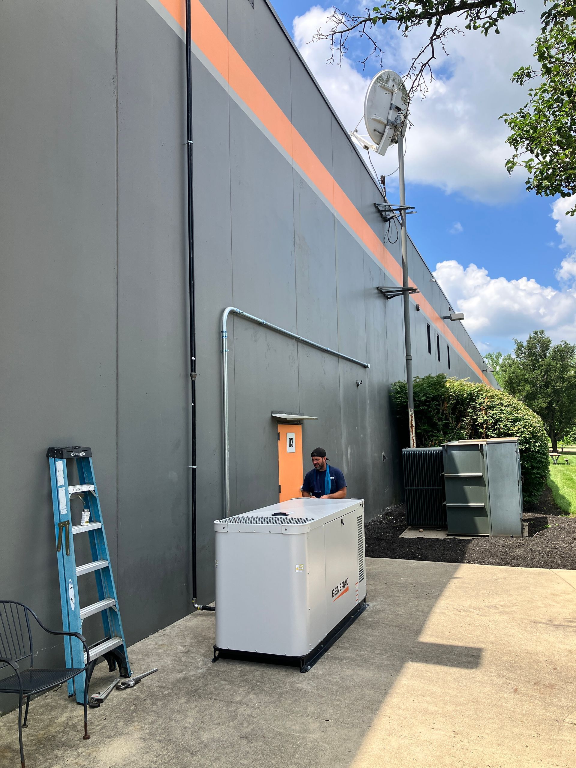 Man near generator on grey building exterior with antennas, orange stripe, and ladder.