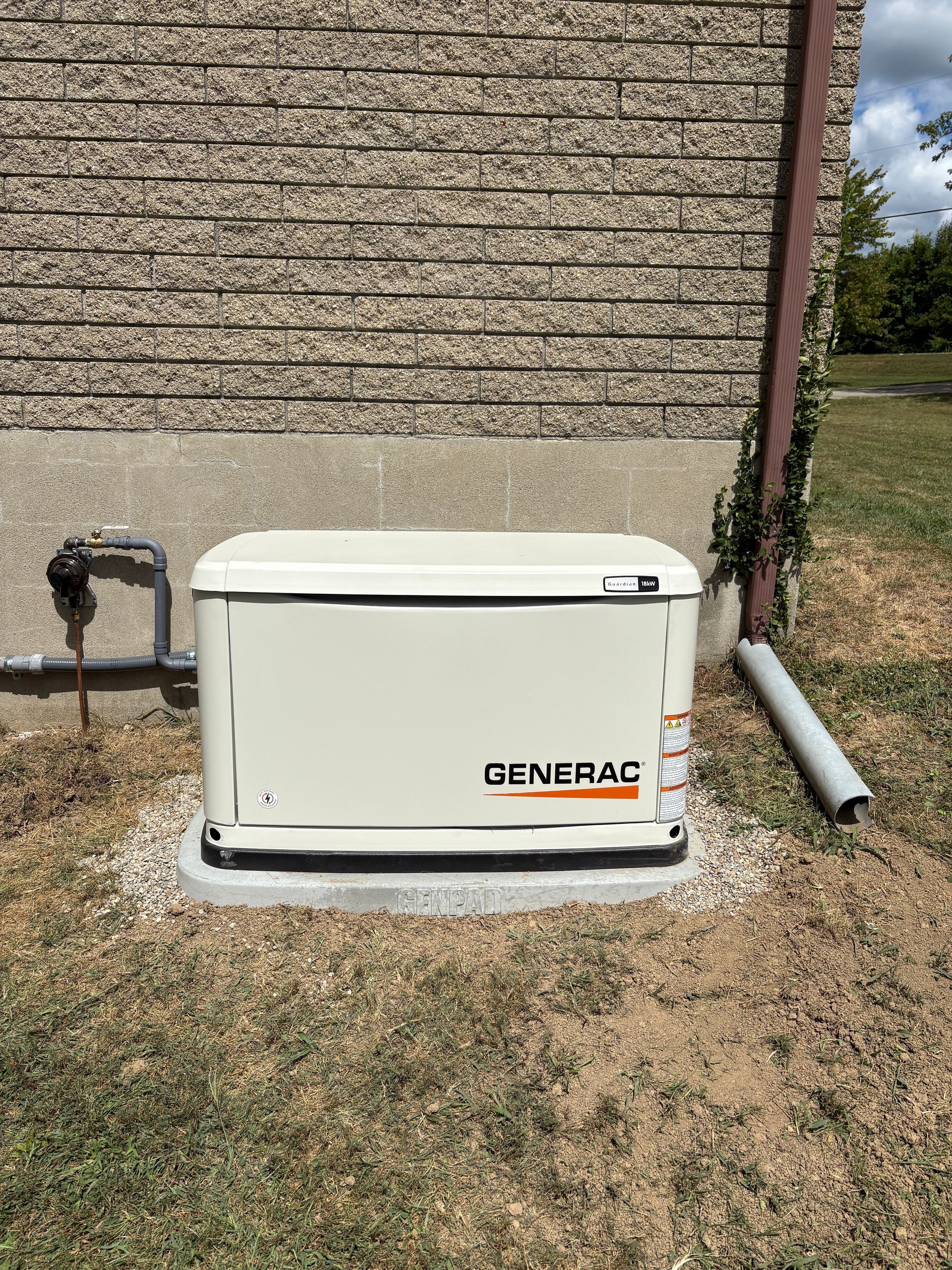 A Generac standby generator, beige, outside a brick building on gravel, with a pipe next to it.