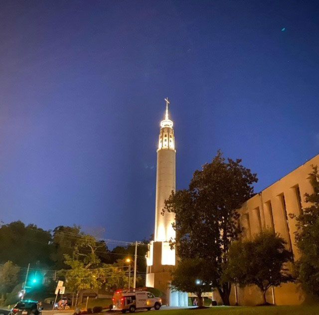 Temple illuminated at night under a dark blue sky with a cross on top and a service vehicle in the foreground.