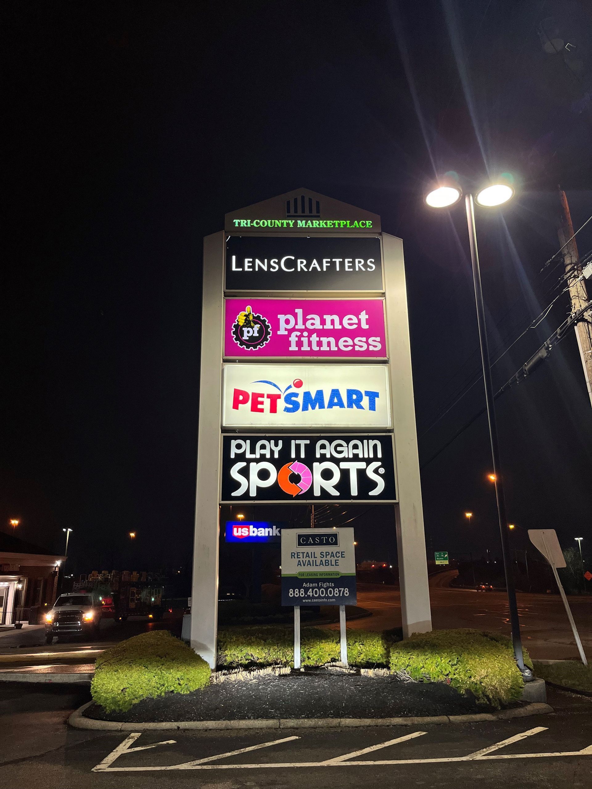 Sign for shopping plaza with stores: LensCrafters, Planet Fitness, PetSmart, Play It Again Sports. At night.