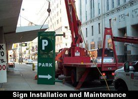 A crane installs a green parking sign in a city, alongside a building and parked vehicles.