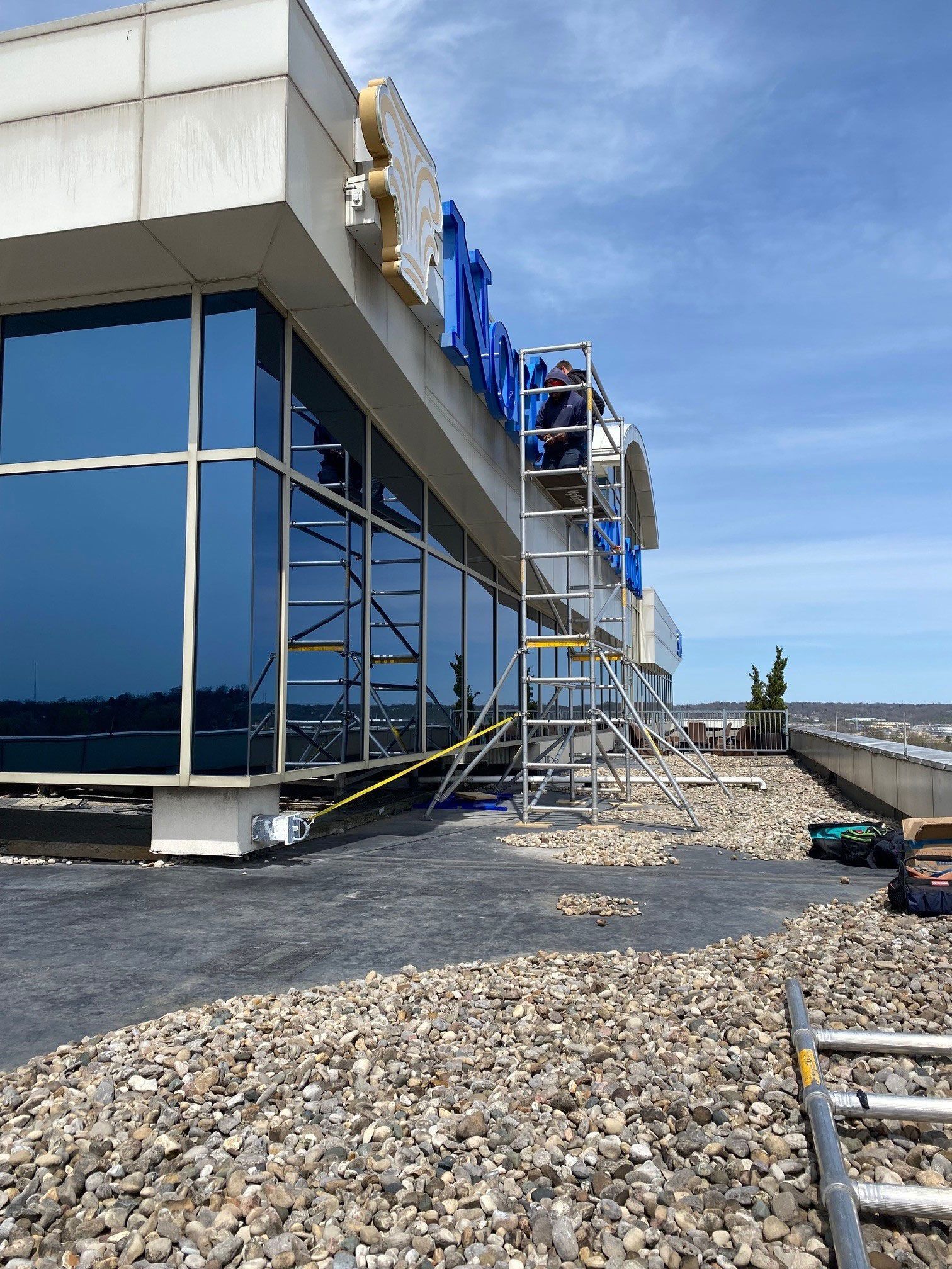 Person on a scaffold working on a building's signage. Exterior shot with blue glass windows and a pebble-covered roof.