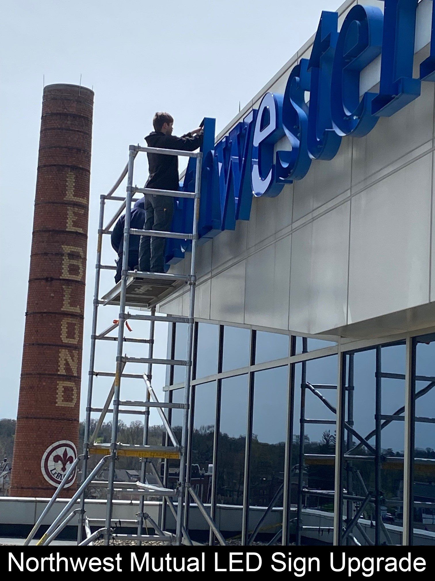 Workers installing LED sign on building; brick smokestack in background.
