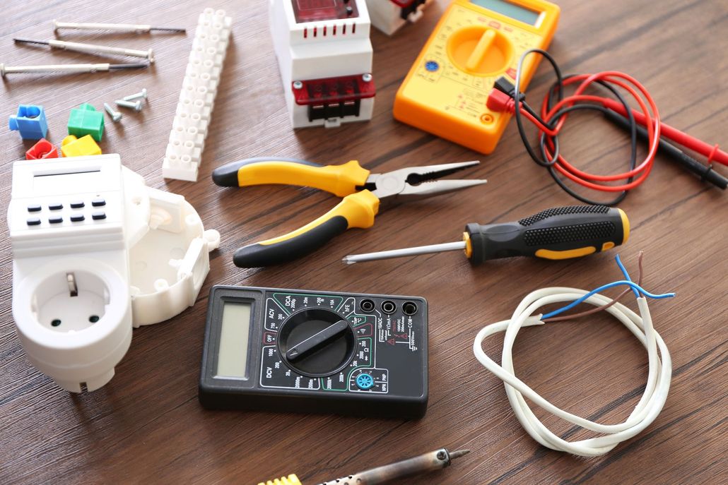 Electrical tools and parts on a table, including a multimeter, pliers, wire, plugs, and cables.