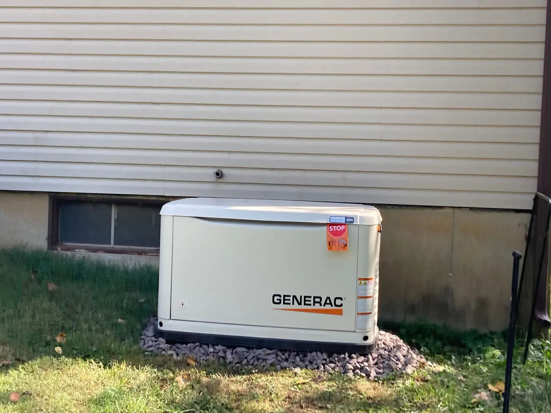 White Generac generator beside a house foundation under beige siding, with grass and gravel around it