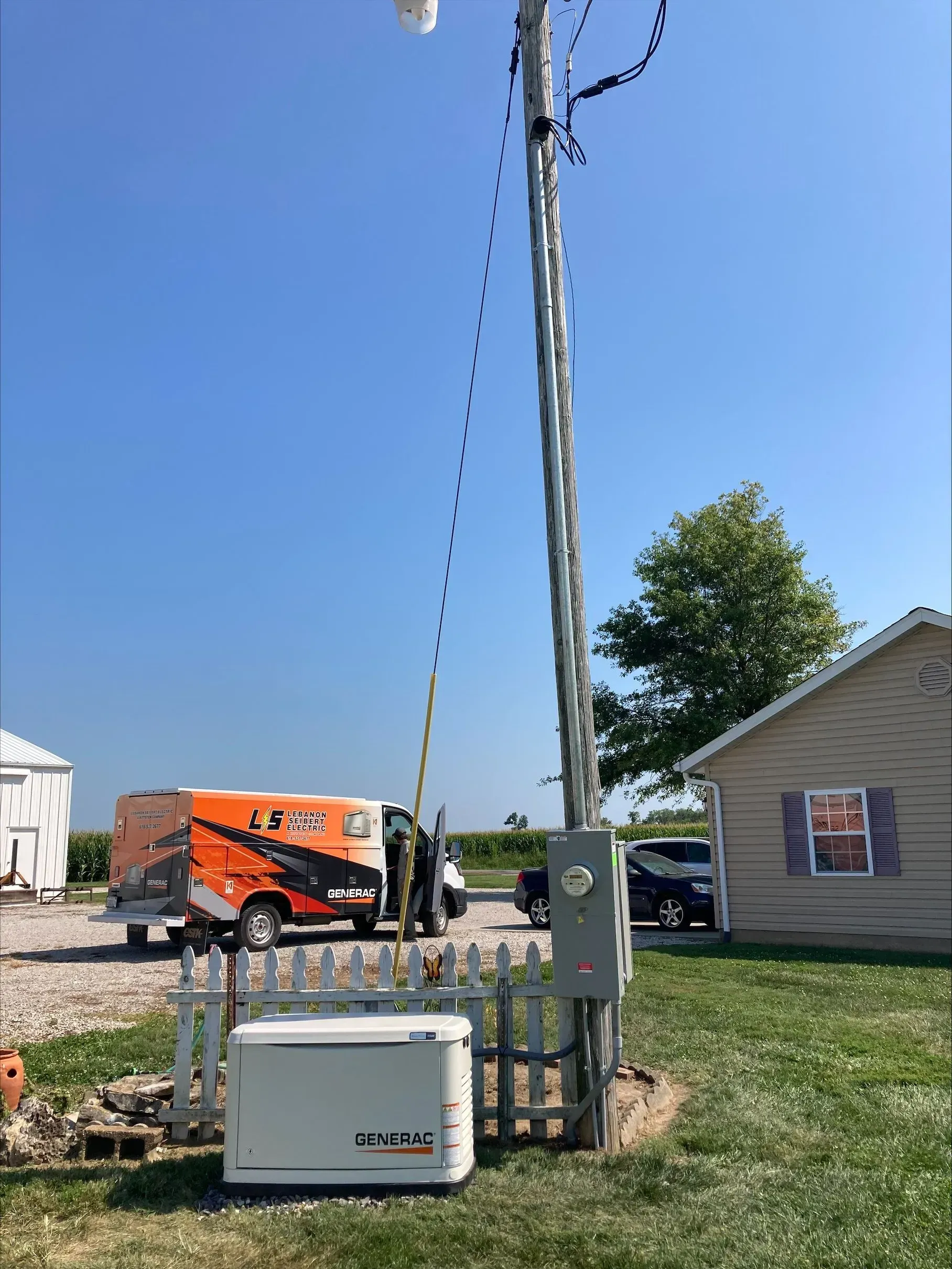 Orange utility truck and workers near a utility pole in a suburban yard, with a house and clear blue sky.