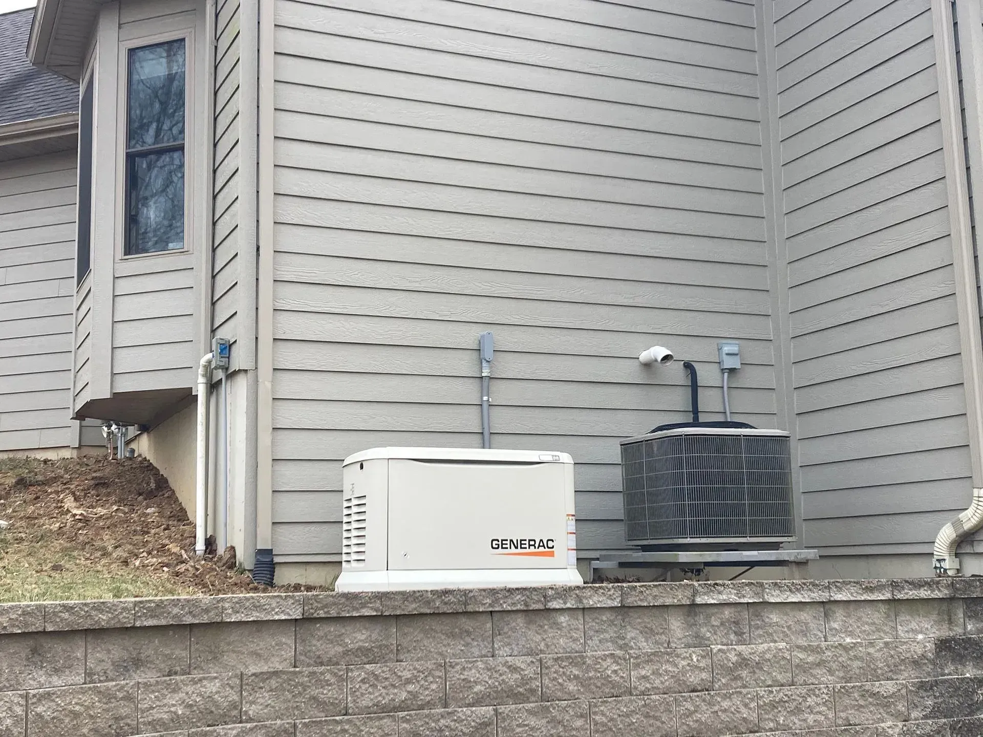 Two outdoor HVAC units beside a house with gray siding and a stone retaining wall