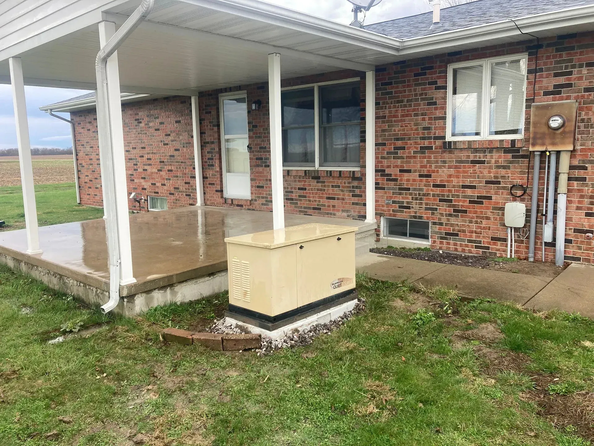 Brick house patio with white posts, wet concrete walkway, and a beige utility box in the yard