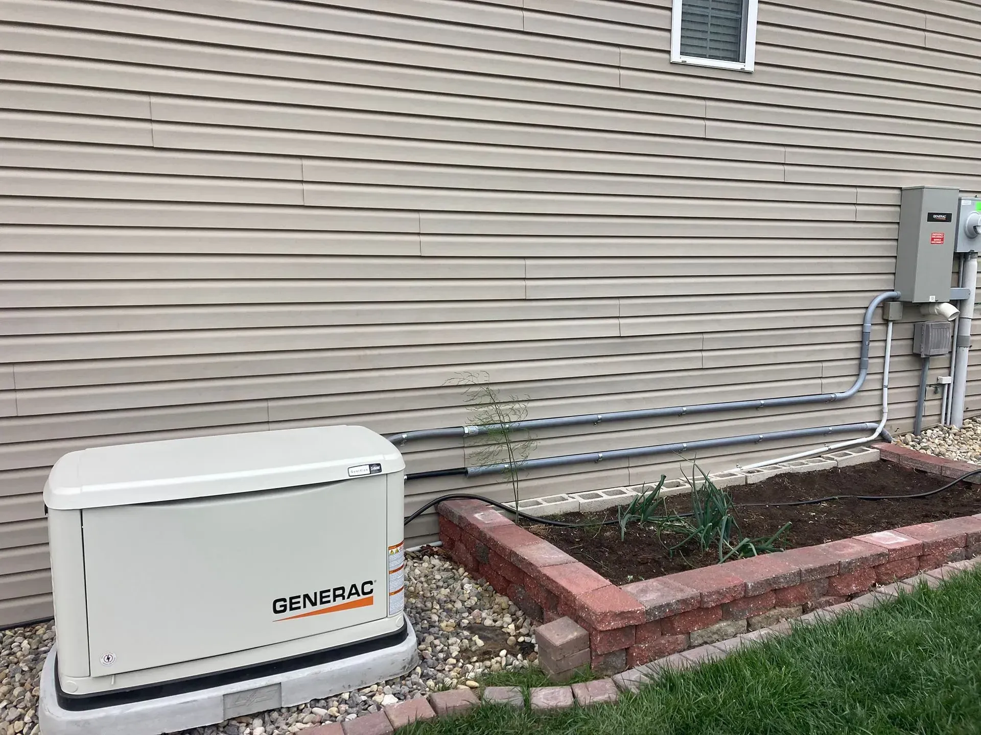 Outdoor utility equipment beside a house wall, with gray conduit pipes and a red brick planter bed.