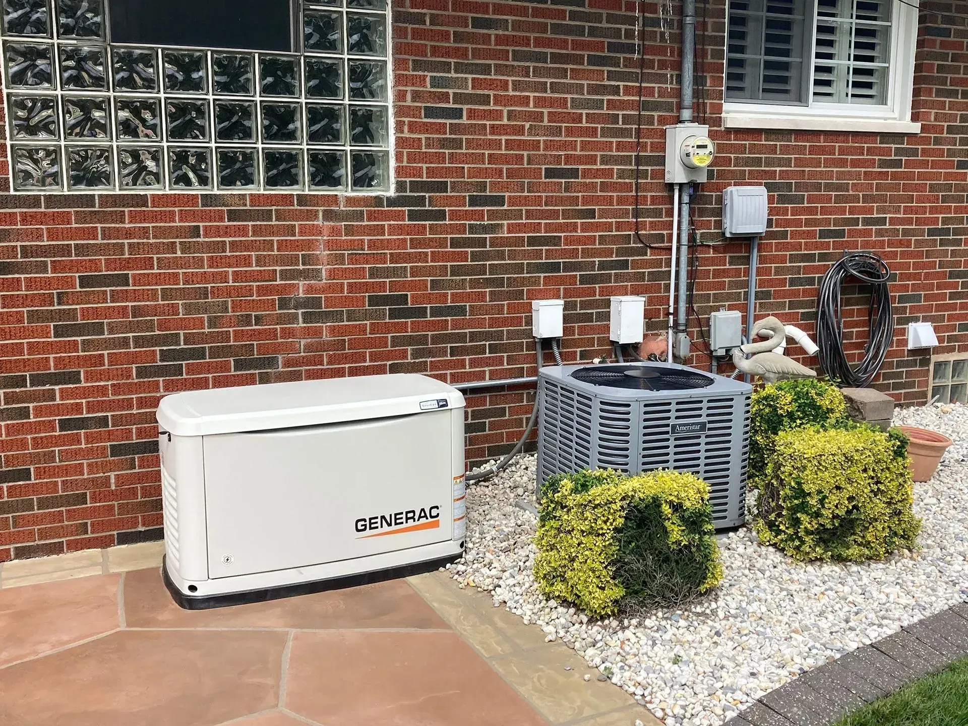 Outdoor generator beside brick house and air-conditioning unit on gravel pad with shrubs.