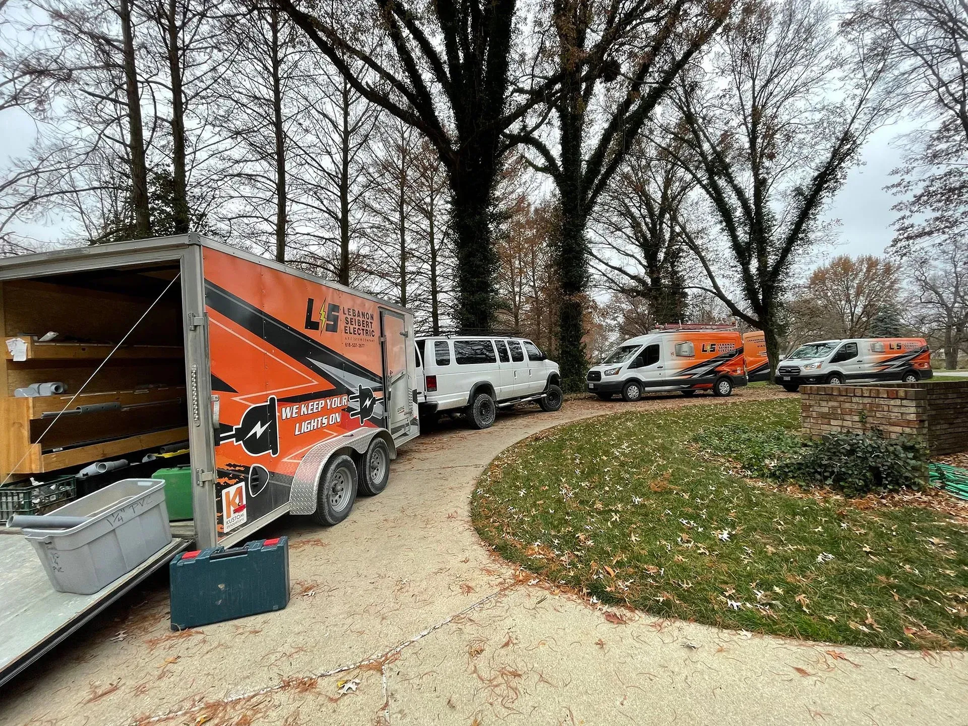 Moving trucks and vans lined up on a driveway beside a house in a wooded yard