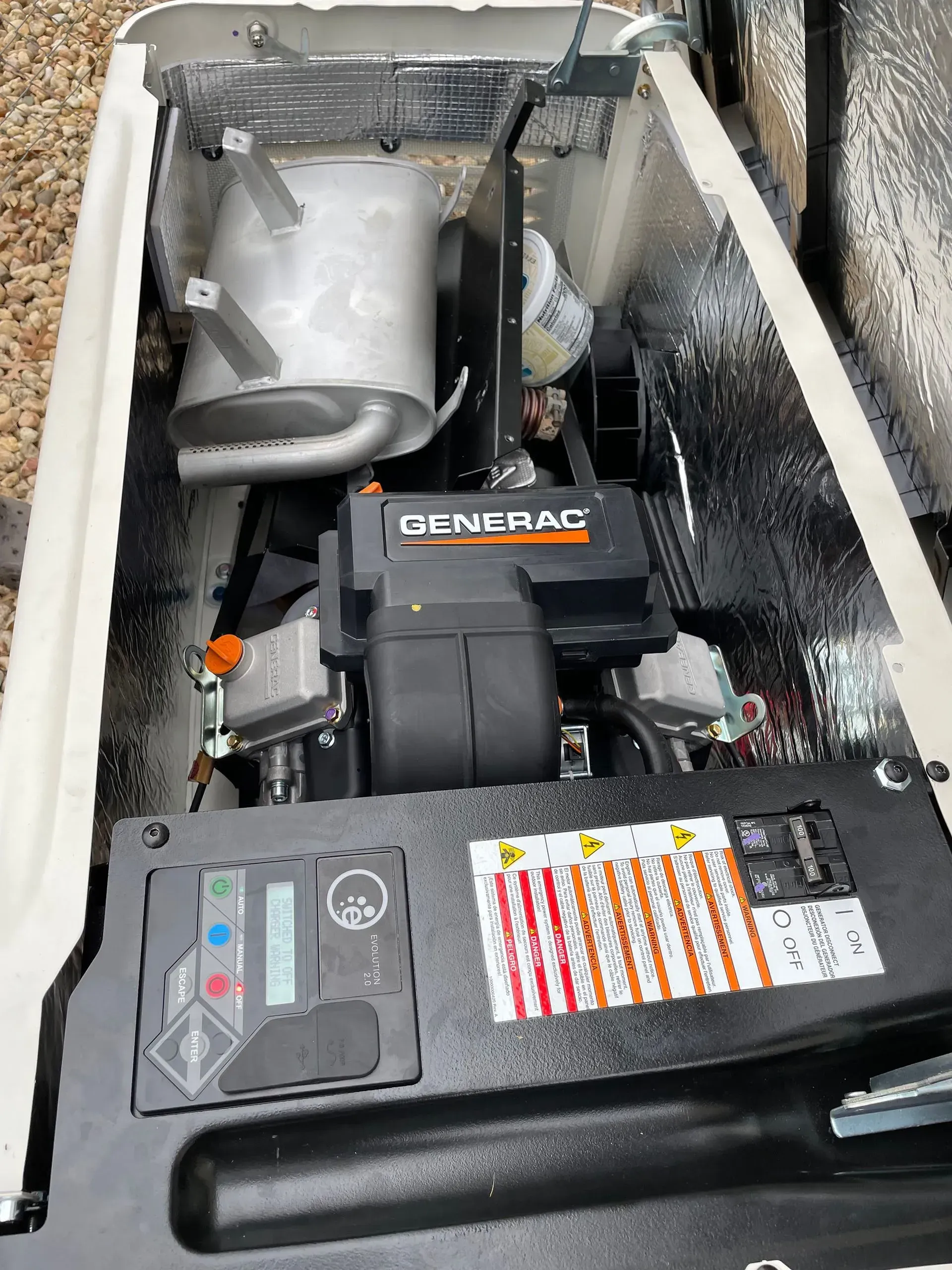 Generator and plumbing equipment in a narrow utility compartment with pipes, tank, and warning labels.