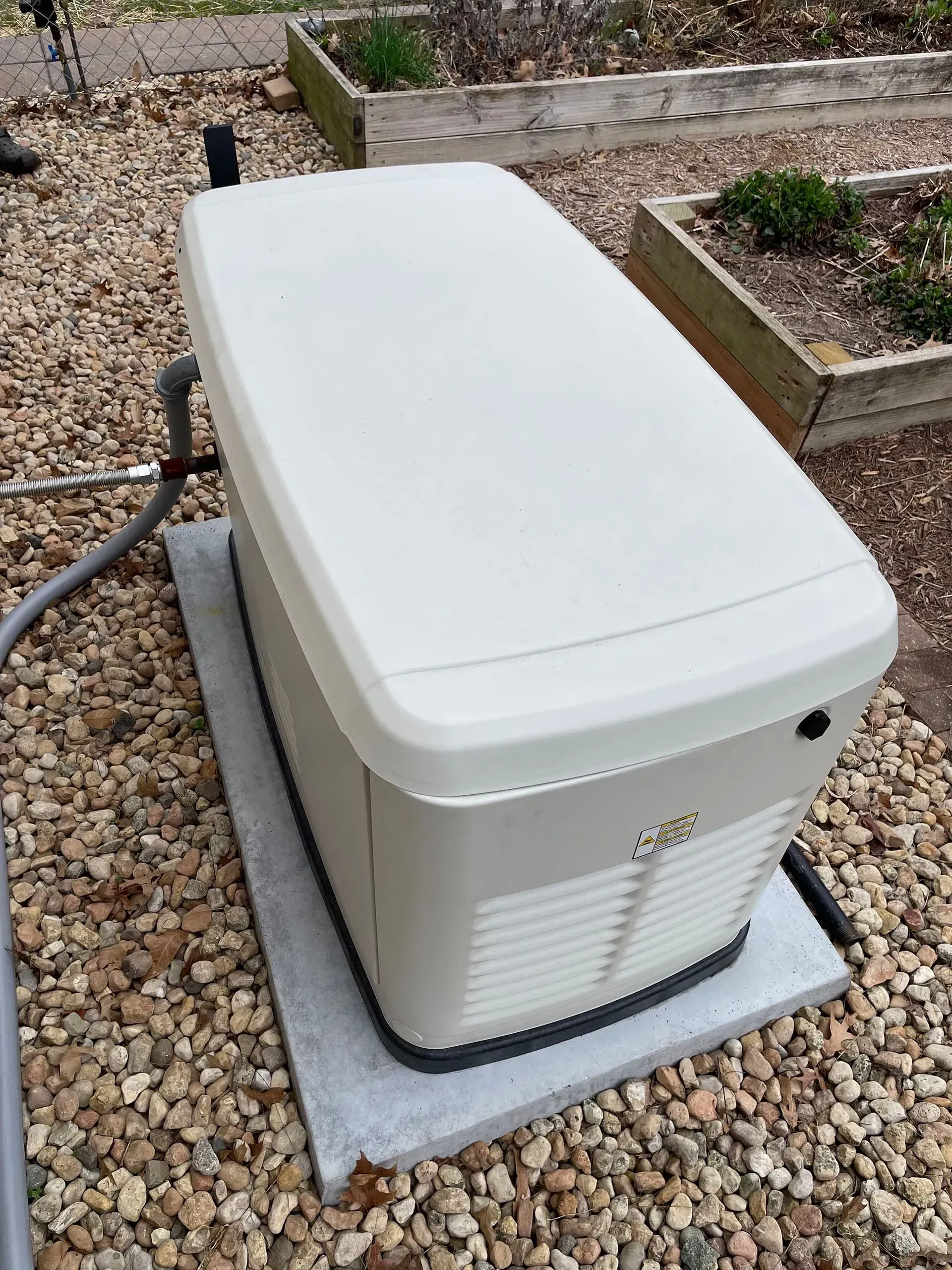 White outdoor generator unit on a concrete pad beside gravel and raised garden beds.