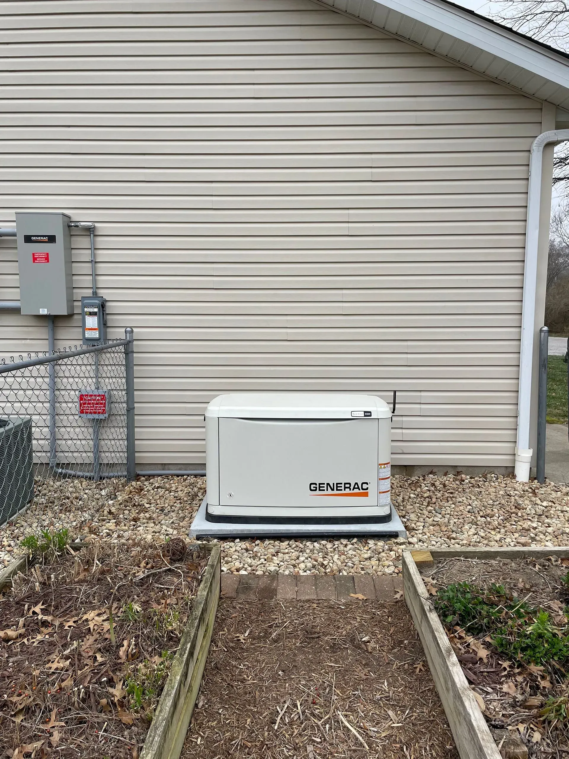 Outdoor generator unit beside a house, with utility meters and a gravel path in front.