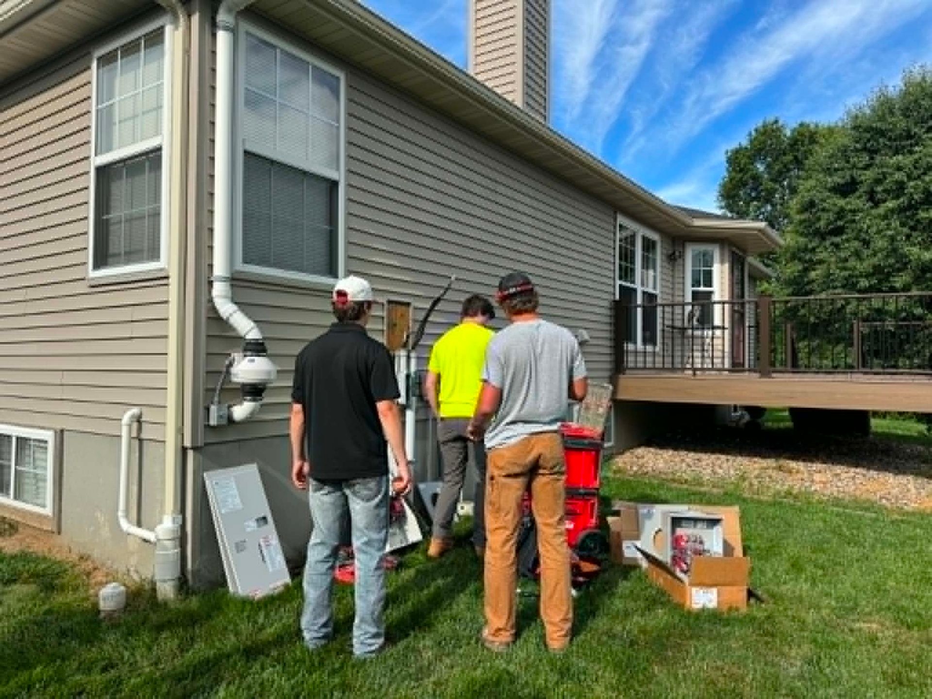 Three workers inspect a house exterior beside a raised deck, with tools and supplies on the grass.