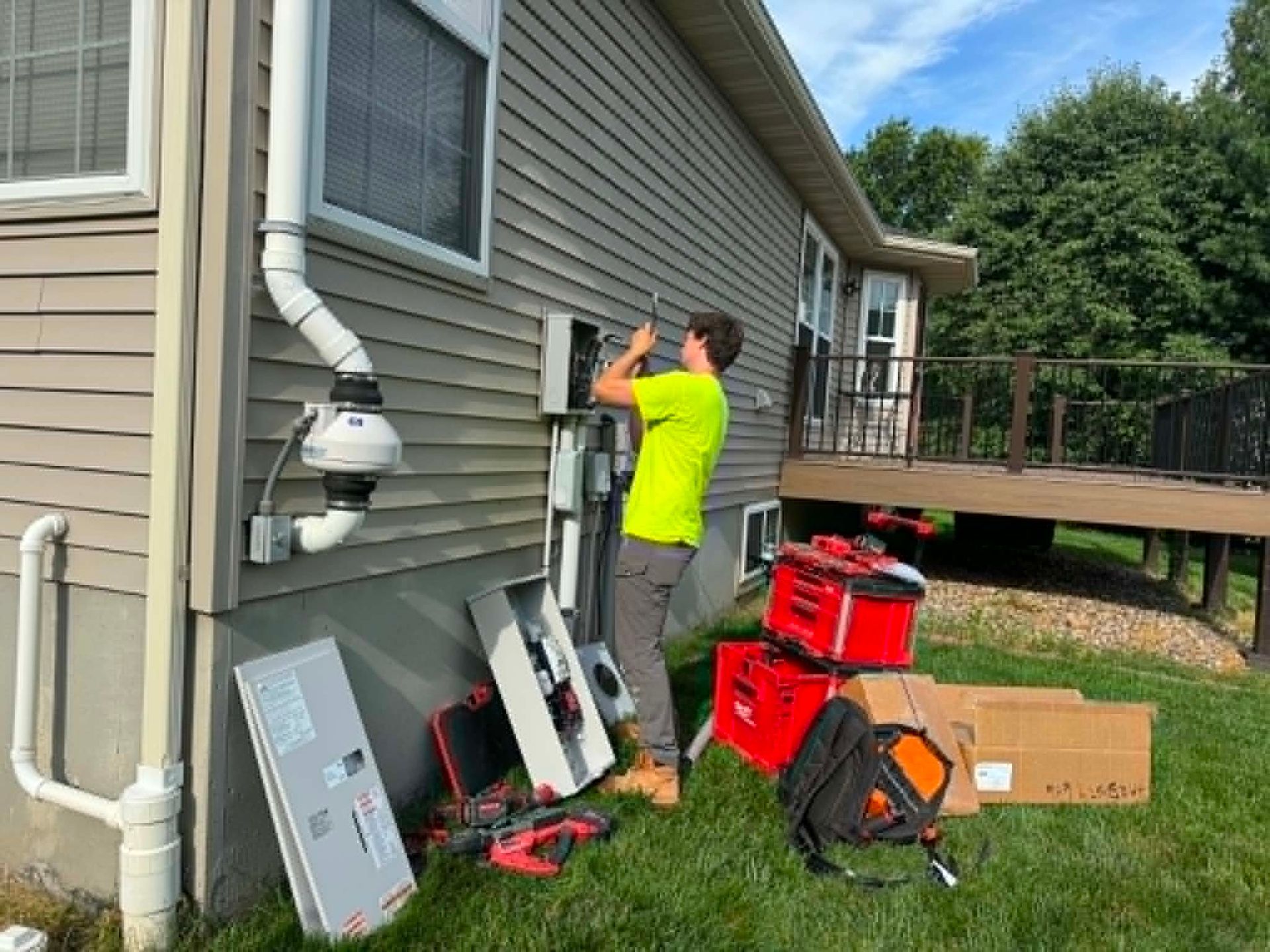 Worker in neon shirt inspecting an outdoor utility panel beside a house, with tools on the ground.