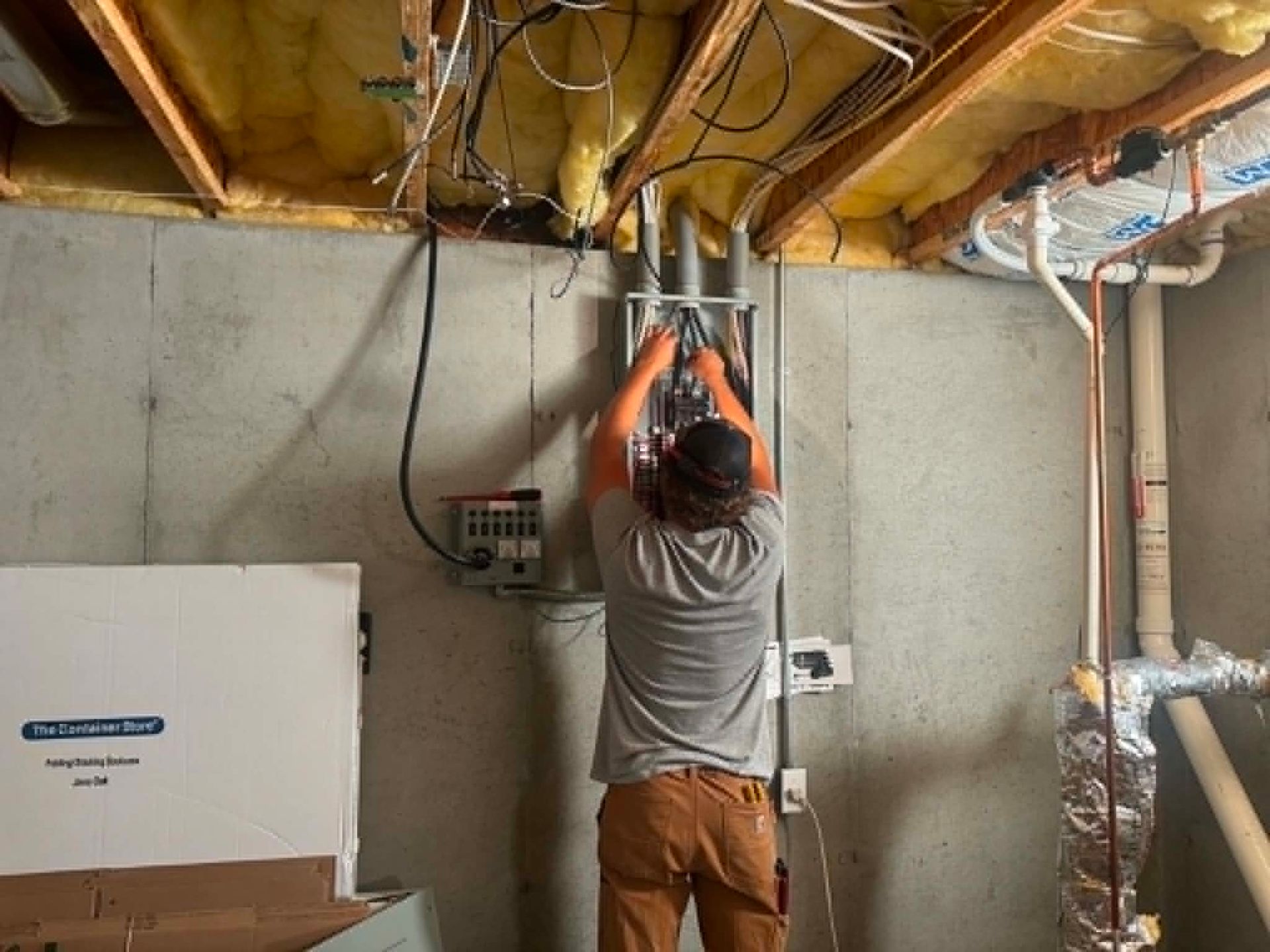 Technician working overhead in an unfinished basement, adjusting wiring near exposed insulation and ductwork.