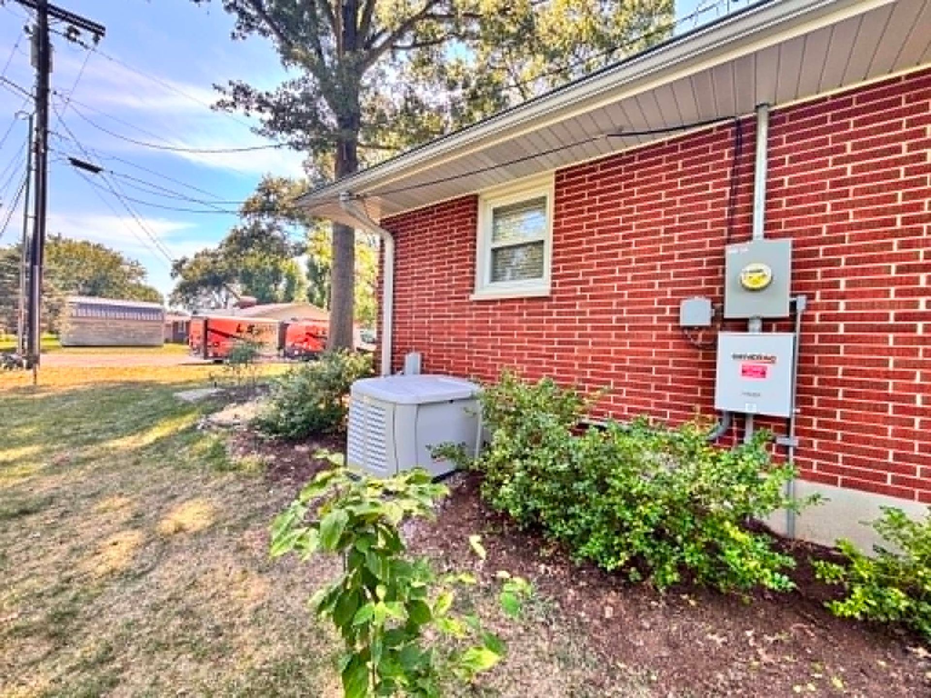 Side of a red brick house with utility meters, HVAC unit, and shrubs along a grassy yard.