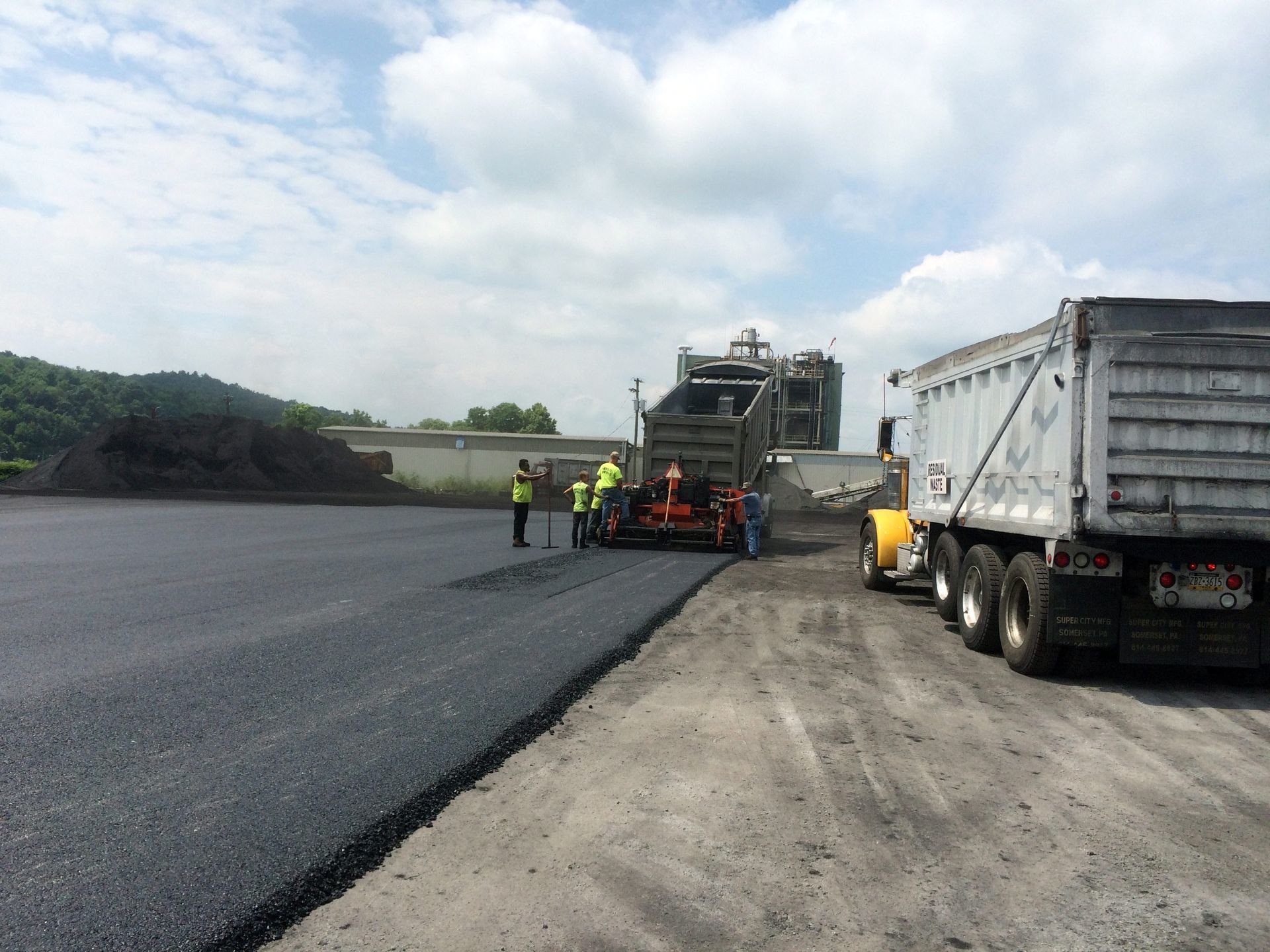 Asphalt paving in progress: workers, asphalt paver, dump truck, and dark asphalt on a grey surface. 