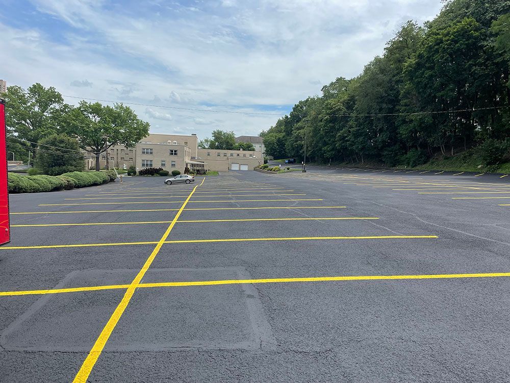Empty asphalt parking lot with yellow lines, trees, and buildings under a cloudy sky.