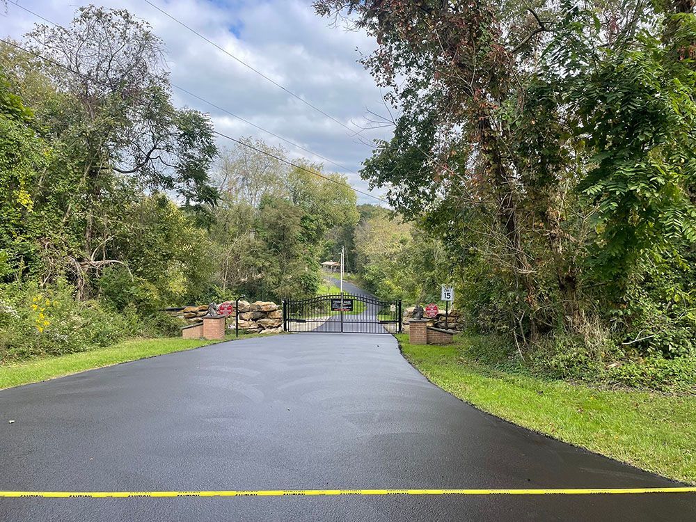 Paved road blocked by gate, trees line sides, green foliage, cloudy sky.
