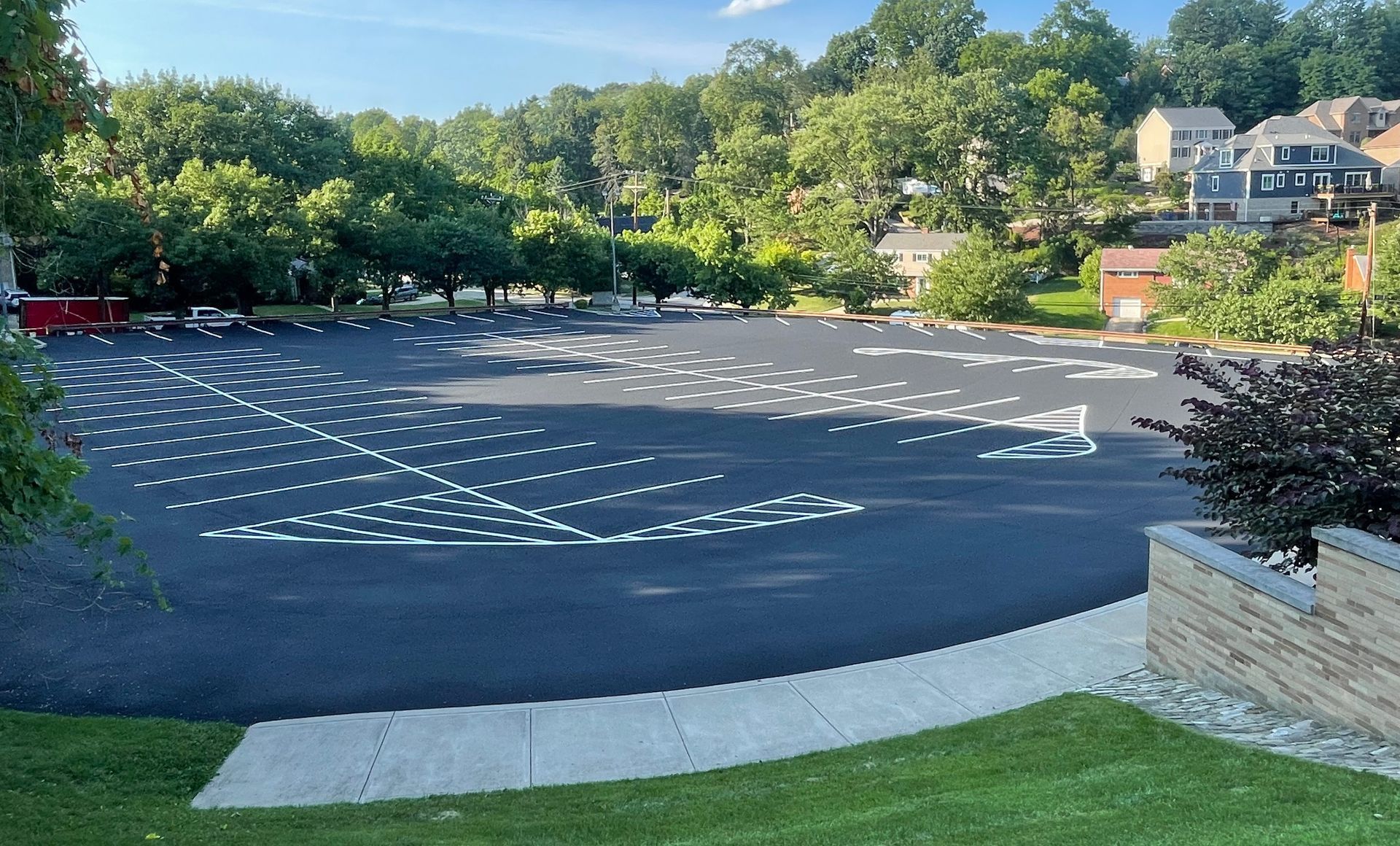 An empty asphalt parking lot with white painted parking spaces, surrounded by trees and a few houses on a hill. 