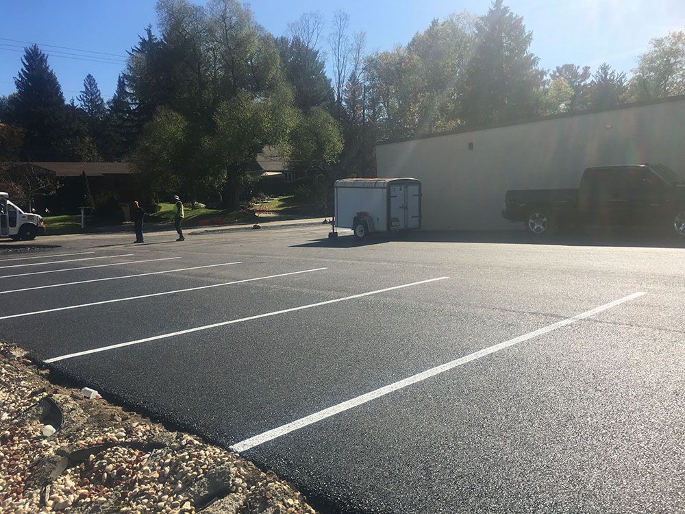 Freshly paved parking lot with white lines, trailer, and trees in background.