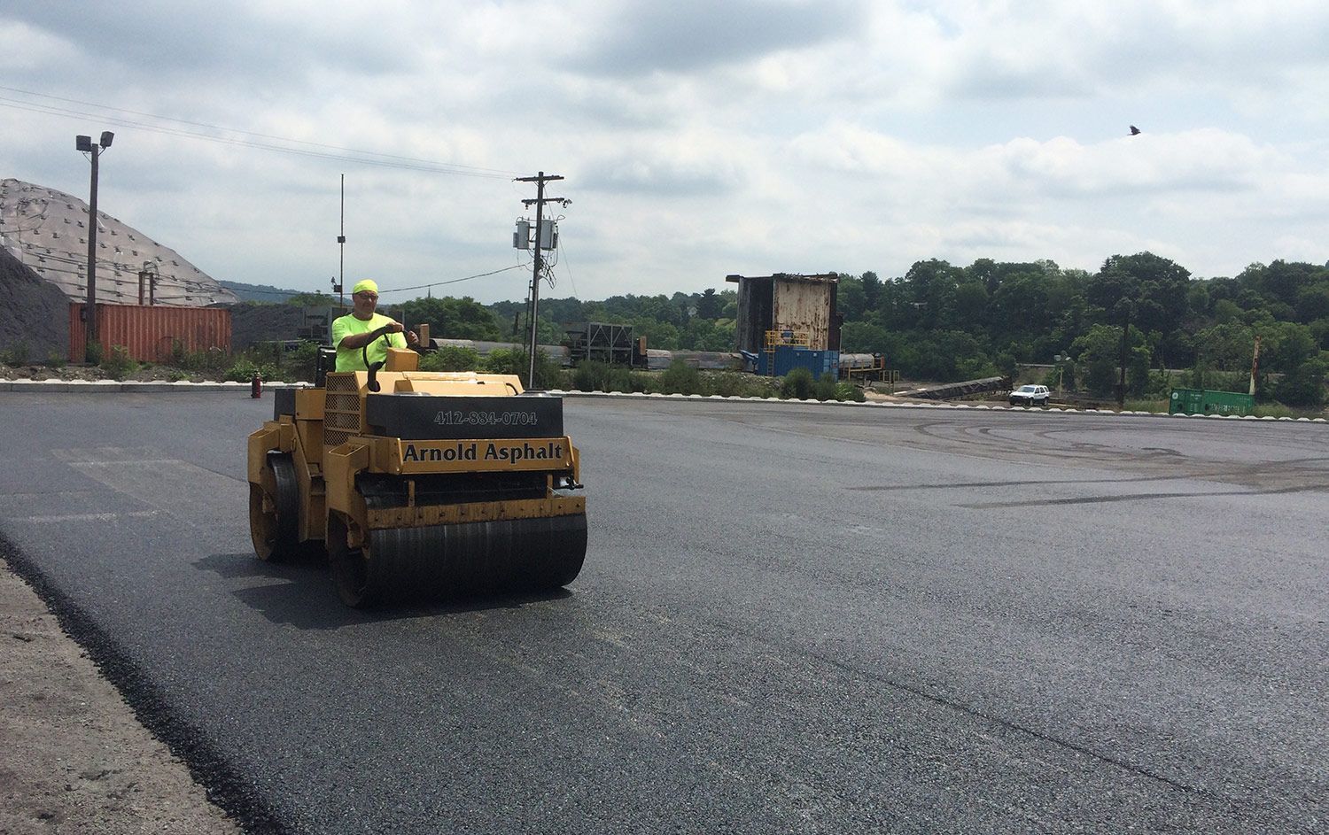 A construction worker operates a yellow asphalt roller on a freshly paved lot under a cloudy sky. 