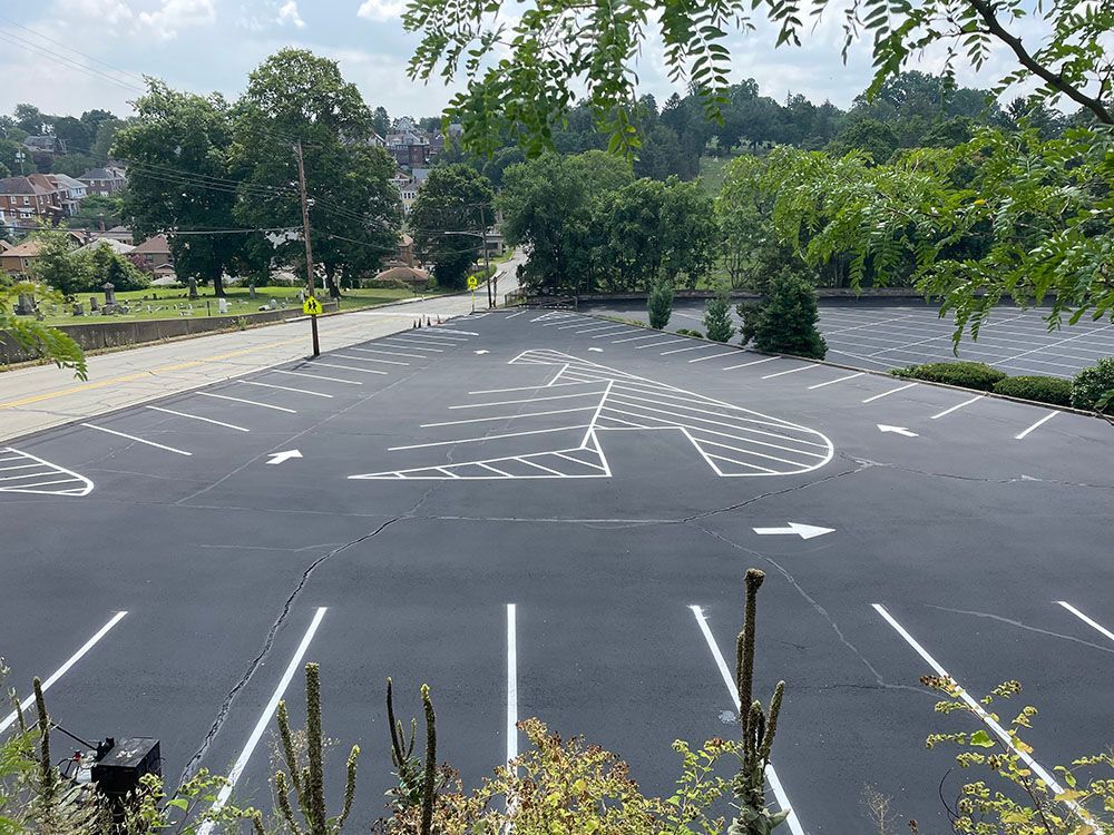 Empty asphalt parking lot with white painted lines and a small basketball court design, surrounded by trees.