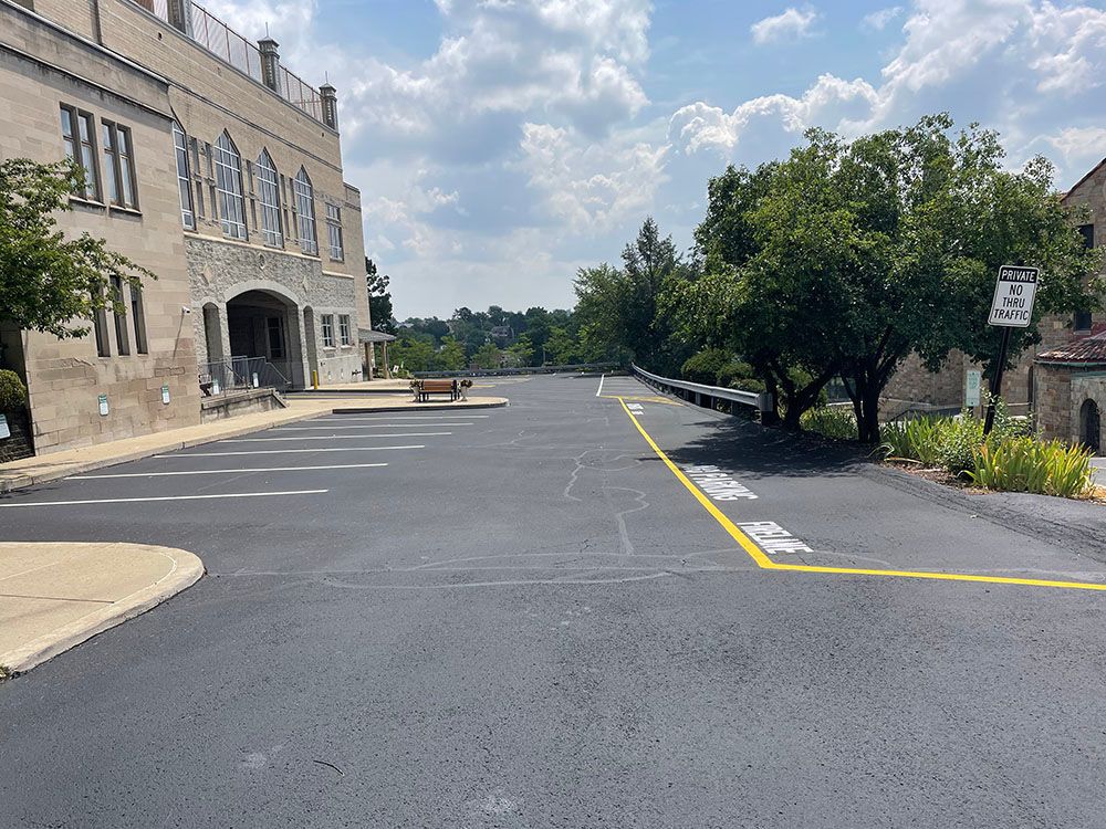 Empty parking lot with freshly paved asphalt, next to a large stone building and trees.