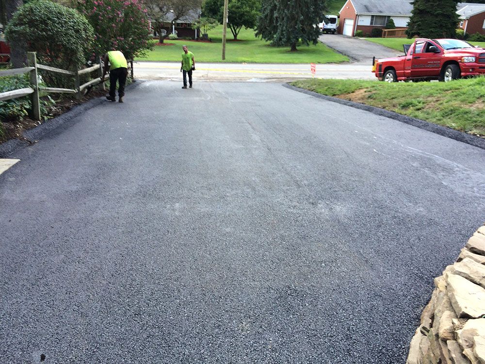 Two workers on a newly paved driveway. One is sweeping; a red truck sits nearby.