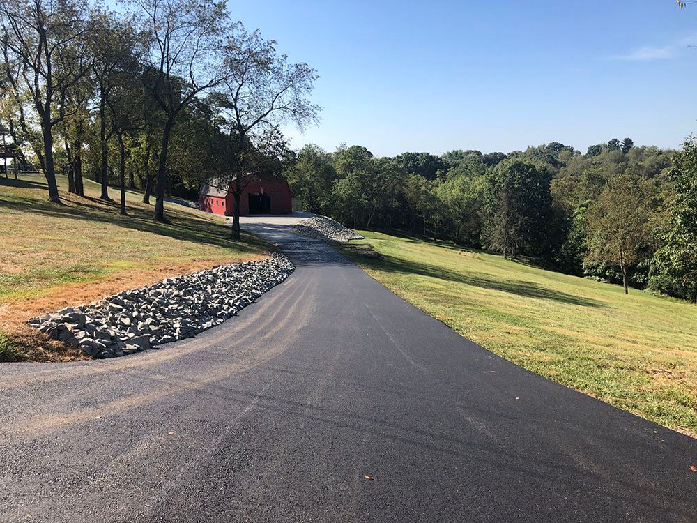 Asphalt driveway leading uphill to a red barn in a green, grassy field.