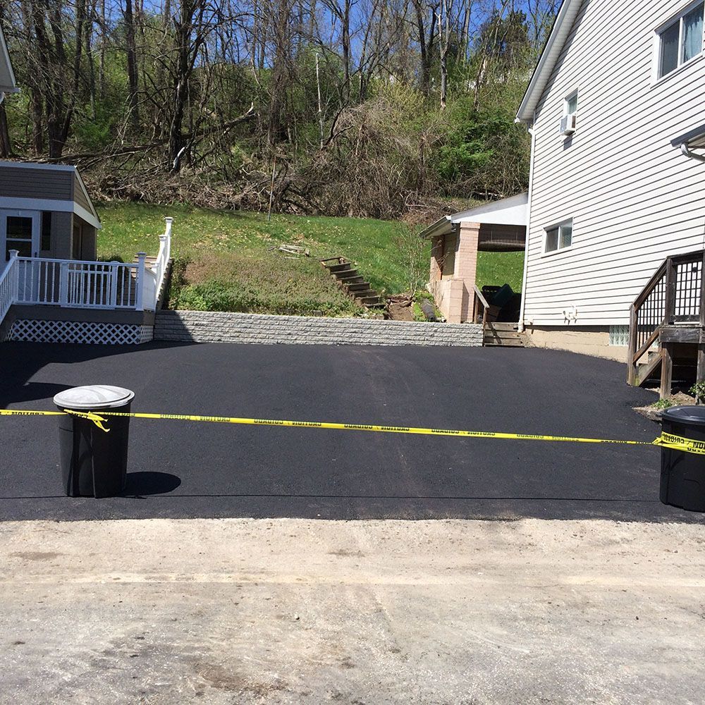Newly paved black driveway with caution tape, two trash cans, and two houses.