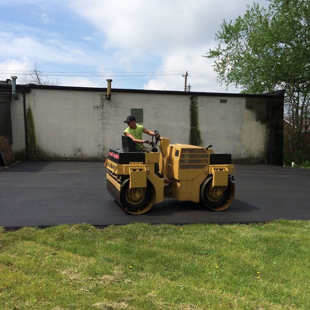Person operating a yellow asphalt roller on a freshly paved parking lot. Green grass surrounds.