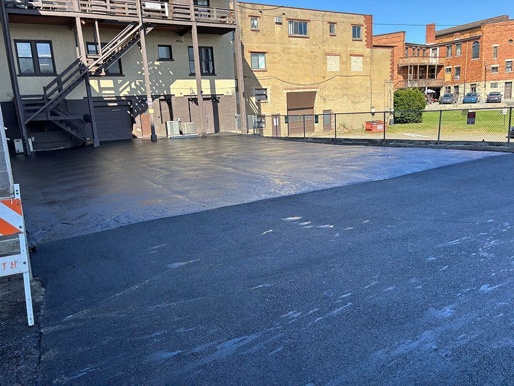 Newly paved asphalt lot with surrounding buildings and a small fence.