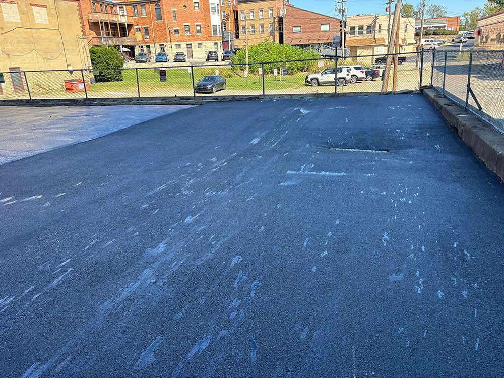 Newly paved asphalt parking lot, light streaks, with buildings and cars visible in the background.