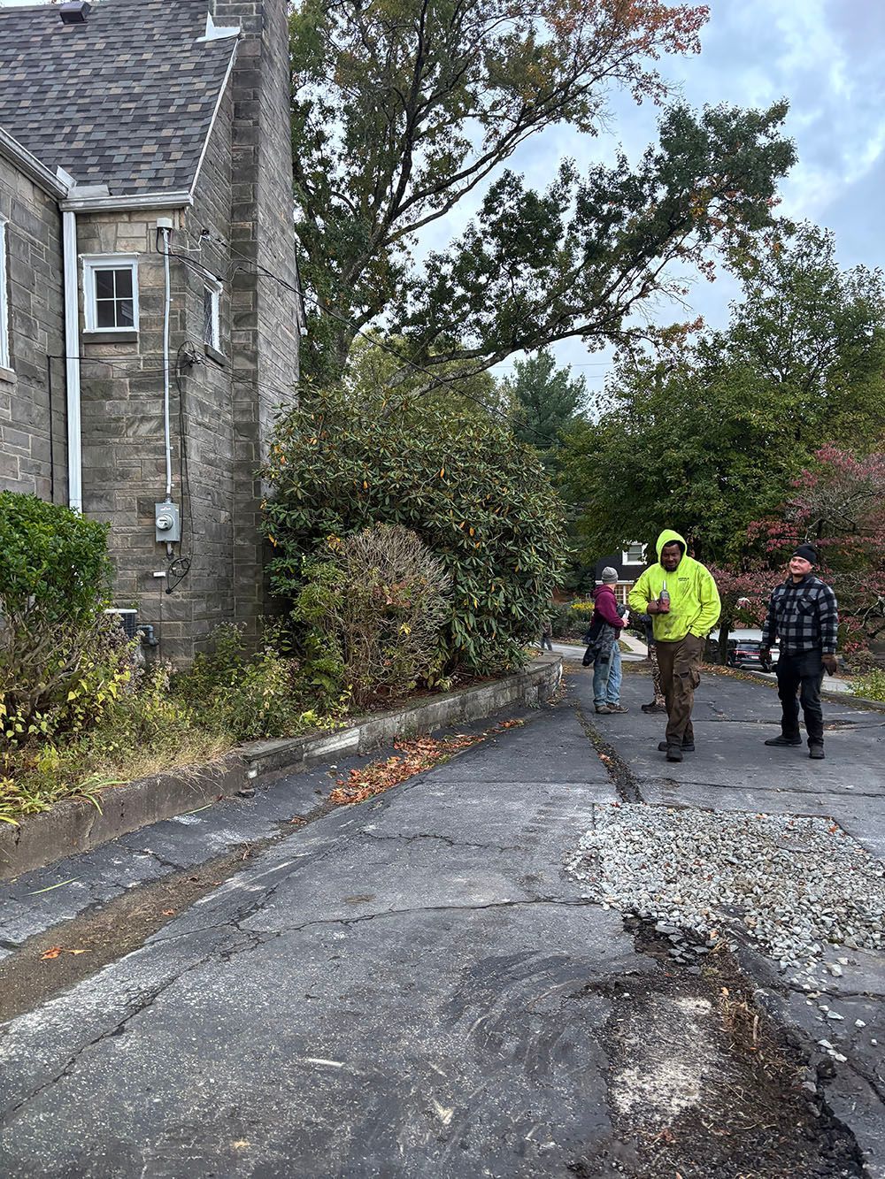 People standing on a cracked asphalt driveway next to a stone building; some foliage.