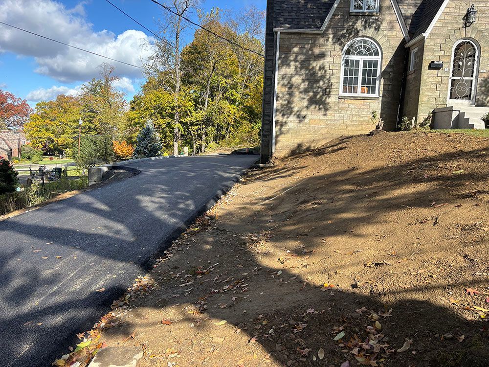 Asphalt driveway next to a dirt embankment and stone house. Fall foliage in the background.