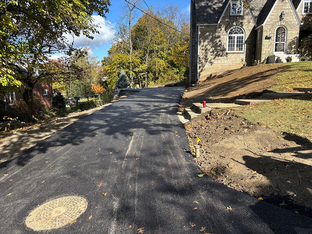 Newly paved asphalt driveway with dirt and grass alongside a stone house on a sunny day.