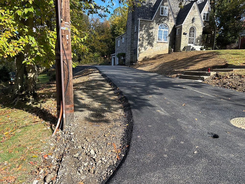 Asphalt driveway next to a dirt shoulder. Utility pole on left, stone house in background. Sunny day.