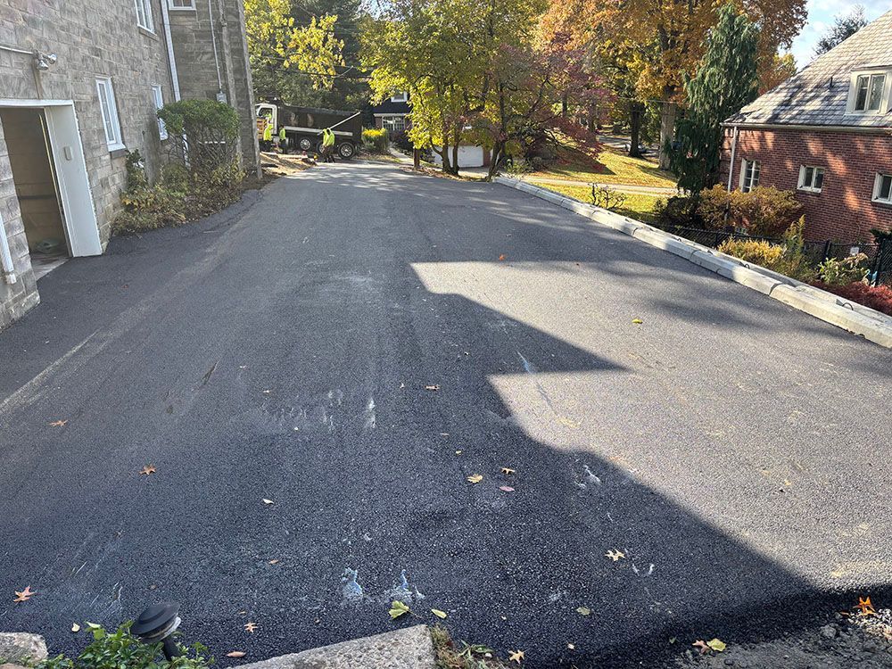 Newly paved asphalt driveway next to a stone building with trees and a house in the background.