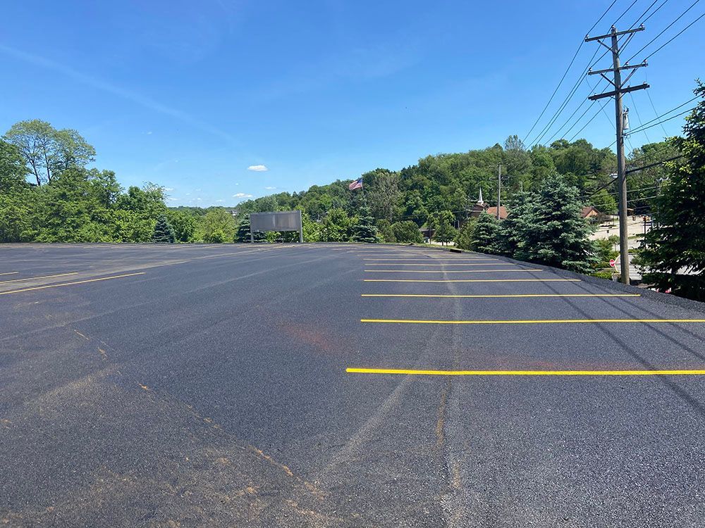 Empty asphalt parking lot with yellow parking lines, trees and a hill in the background under a blue sky.