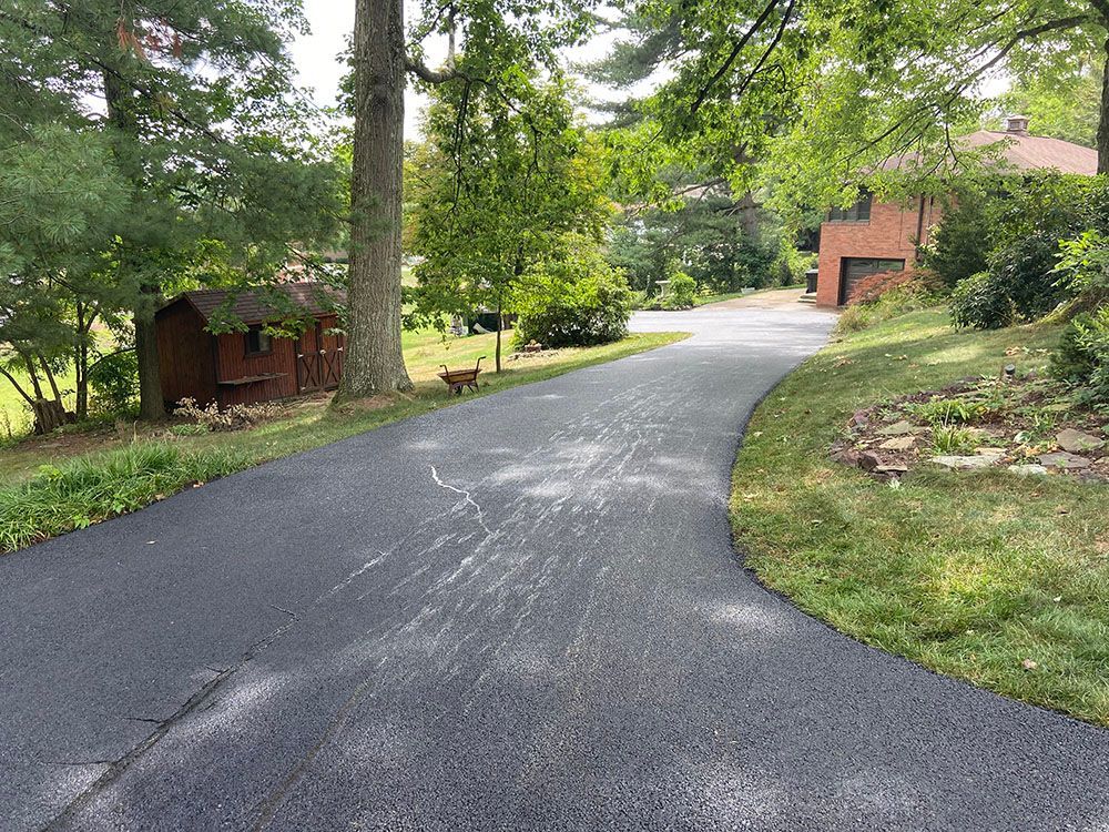 Paved driveway curves uphill past a shed, trees, and a brick house. White streaks on asphalt.